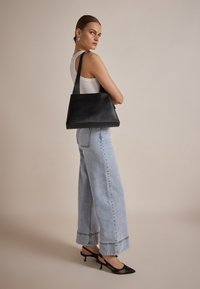 Schwarze Lederhandtasche in dreieckiger Form, mit flachem Boden und breiten Trägern. Das Model trägt hellblaue weit geschnittene Jeans und schwarze Absätze.