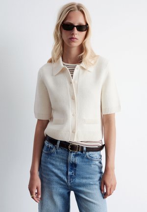 White short-sleeve cardigan with a collar and button closure, featuring two front pockets. Worn over a striped shirt and blue jeans.