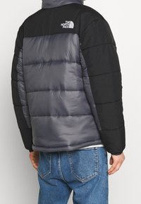 Polstret jakke med en grå krop og sorte ærmer samt et logo på den øverste del af ryggen. Materialet ser glat ud med en let blank finish.