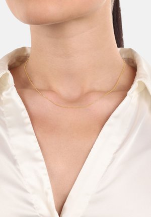 Collana color oro con una delicata catena sottile che riposa su un collo nudo, abbinata a una blusa bianca setosa con scollo a V.