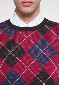 Pull en argyle rouge et marine avec un col côtelé noir, featuring des coutures blanches et un petit logo rouge en bas à gauche. Chemise blanche à col en dessous.