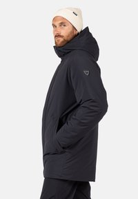 Chaqueta aislante oscura con capucha, que cuenta con bolsillos laterales y un logo en la manga. Llevado con un gorro de punto beige claro.