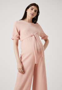 Rosa teksturert graviditets jumpsuit med korte pufferm og knytning i midjen. Løs passform med vide bein i en subtil krøllet stoff.