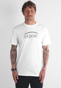 Homme avec des tatouages sur les deux bras portant un t-shirt blanc avec le logo OXBOW et un pantalon noir, se tenant devant un fond clair.