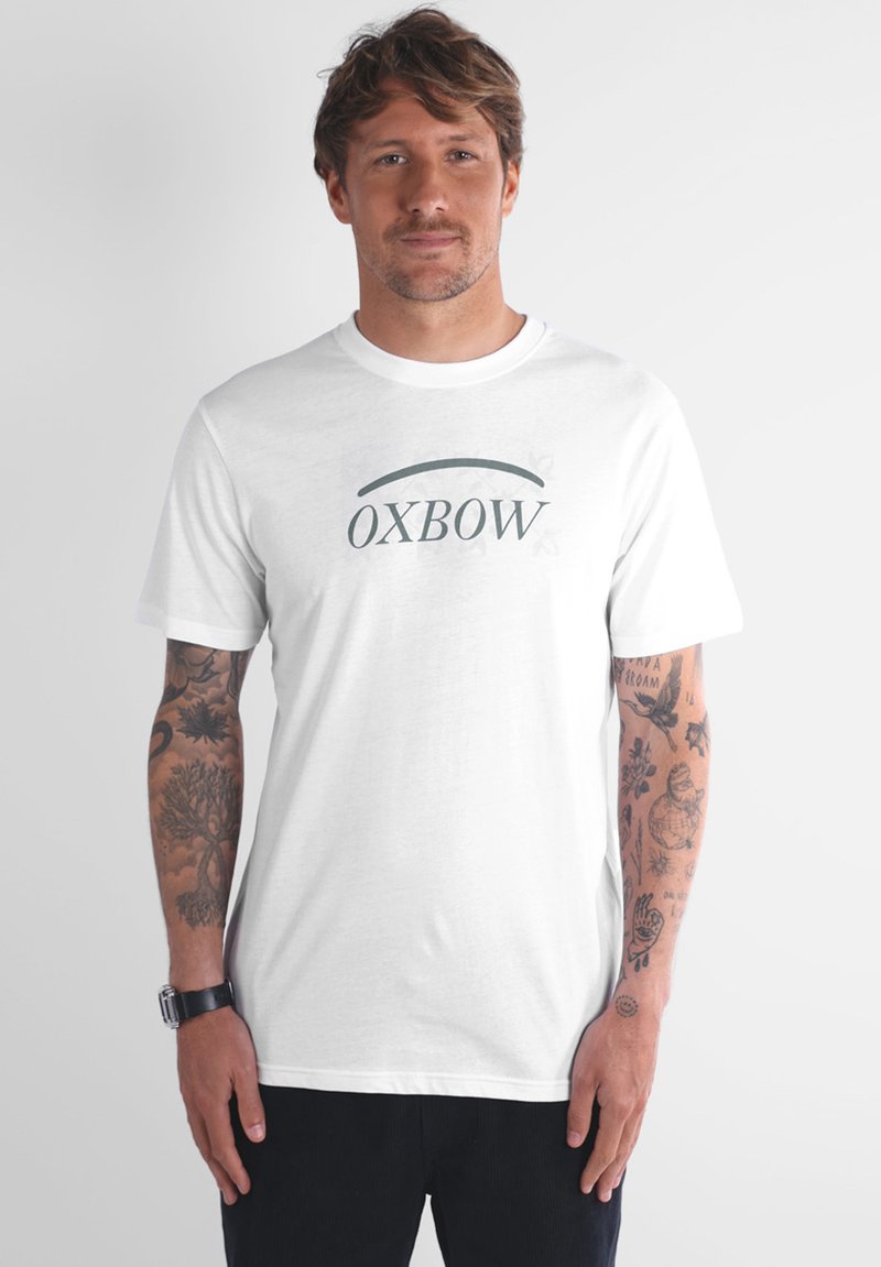 Homme avec des tatouages sur les deux bras portant un t-shirt blanc avec le logo OXBOW et un pantalon noir, se tenant devant un fond clair.