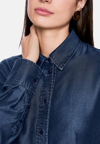 Dark blue denim shirt with a button-up front, pointed collar, and long sleeves. Features smooth texture and subtle sheen.