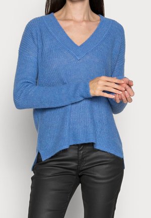 Kobieta ubrana w niebieski sweter z dekoltem w serek, z długimi rękawami, oraz czarne skórzane spodnie na jednolitym tle.