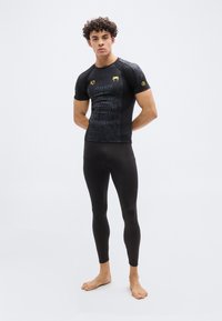 Νεαρός άνδρας με σγουρά μαλλιά, φορώντας εφαρμοστό μαύρο αθλητικό μπλουζάκι με χρυσά λογότυπα και μαύρο κολάν, όρθιος ξυπόλητος μπροστά σε μονόχρωμο φόντο.