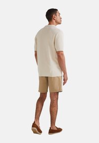 Pull court beige clair à manches courtes avec un design en maille texturée, associé à un short kaki et des chaussures brunes à enfiler, vu de derrière.