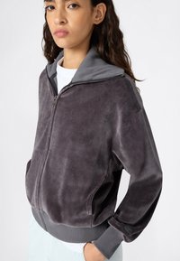 Veste grise zippée avec une texture douce et veloutée, des poignets côtelés et une capuche contrastante. Elle dispose de poches latérales et de détails matériels minimaux.