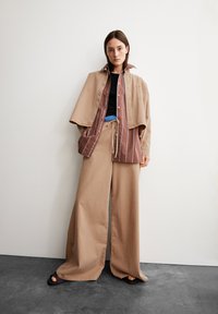 Veste oversize beige superposée sur une chemise rayée marron et blanche. Pantalons à jambes larges beiges et haut noir. Des sandales simples complètent la tenue.