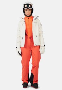 Lyžařský outfit zahrnující bílou voděodolnou bundu, oranžovou spodní vrstvu, oranžové kalhoty s černými akcenty a bílé rukavice. Vybavená helmou a brýlemi.