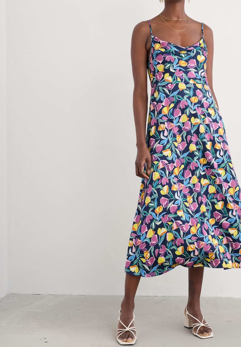 Femme portant une robe midi sans manches bleu marine avec un imprimé floral rose, jaune et bleu, et des sandales blanches à talons carrés à brides.