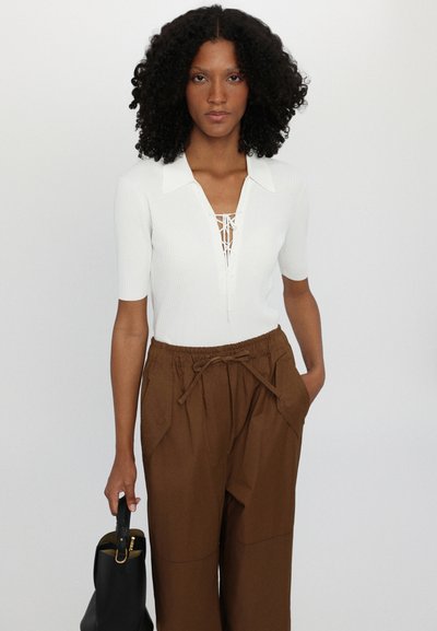 White ribbed short-sleeve top with collar and lace-up front. Paired with brown, loose-fitting, elastic-waist pants and a black handbag.
