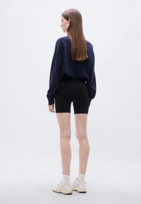 Μπλούζα από βαμβάκι σε ναυτικό μπλε, μαύρο εφαρμοστό σορτς, λευκές κάλτσες μέχρι τους αστραγάλους και μπεζ αθλητικά παπούτσια. Το μοντέλο στέκεται με την πλάτη σε απλό φόντο.