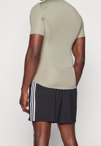 Krótki rękaw sportowa bluzka w jasnooliwkowym kolorze, dopasowany krój; w połączeniu z czarnymi szortami z białymi paskami po bokach.