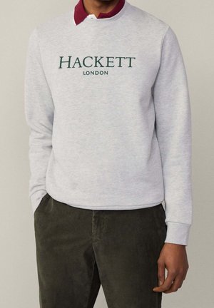 Uomo che indossa una felpa grigio chiaro con la scritta "HACKETT LONDON", una camicia bordeaux con colletto sotto e pantaloni di velluto a coste verde scuro.
