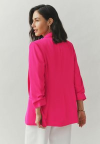 Fuchsiový prispôsobený blejzer z ľahkej tkaniny s výrezom na golieri, zrolovanými rukávmi a uvoľneným strihom, kombinovaný s bielymi nohavicami.