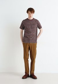 Striped short-sleeve t-shirt in brown with white and navy stripes, paired with brown trousers. Model stands with hands in pockets.
