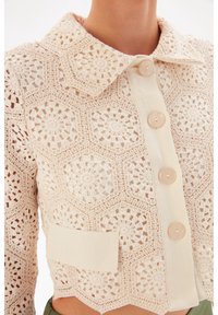 Gehaakte blouse in lichtbeige met hexagonale patronen, een kraag, vijf knopen en een stoffen patch aan de zijkant. Geweven en luchtig ontwerp.