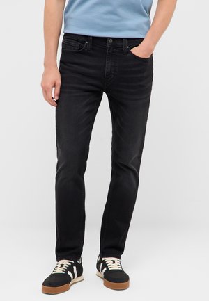 Mann trägt schwarze Slim-Fit-Jeans, blaues T-Shirt und schwarze Sneakers mit weißen Streifen und braunen Sohlen, steht mit einer Hand in der Tasche.