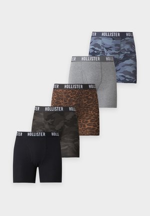 Homme torse nu, debout pieds nus, portant un boxer gris camouflage avec une ceinture à logo Hollister sur fond blanc uni.