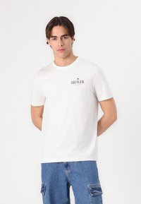 T-shirt bianco in cotone con maniche corte e collo rotondo. Presenta un logo rosso e blu stampato sul petto. Vestibilità casual. Abbinato a pantaloni corti di jeans.