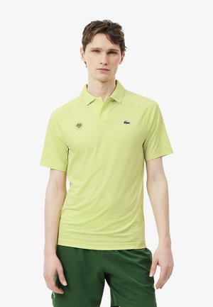 Joven con una camiseta polo Lacoste verde claro y pantalones cortos verde oscuro, de pie frente a un fondo blanco liso.