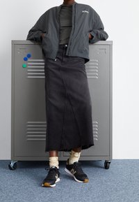 Giacca bomber grigia sopra una maglietta verde scuro, gonna di jeans nera con orlo sfrangiato e sneakers nere con dettagli dorati. Sullo sfondo si trova un armadietto grigio.