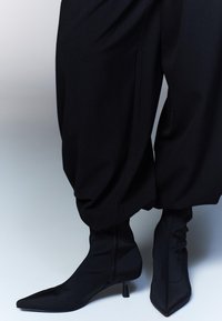 Crne uske čizme s oštrim vrhom i niskom, vitkom petom, napravljene od glatkog, elastičnog materijala, u paru s raskošnim crnim hlačama.