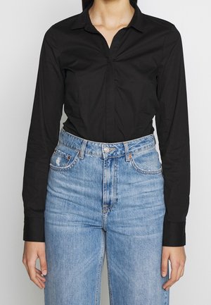 Person iført en tætsiddende sort langærmet skjorte, der er stoppet ned i højtsiddende lyseblå denimjeans med forlommer og bæltestropper.