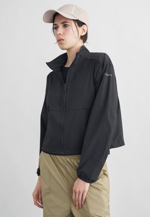 Mlada oseba, oblečena v črno jakno z zadrgo, bež hlače in svetlo bež kapo, stoji pred enakomerno svetlo podlago.