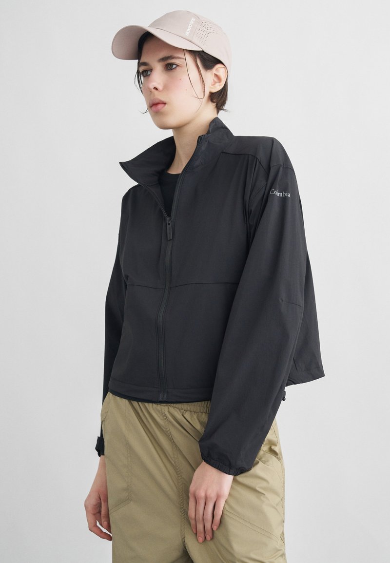 Νεαρό άτομο που φοράει μαύρο μπουφάν με φερμουάρ, μπεζ παντελόνι και ανοιχτό μπεζ καπέλο, στέκεται μπροστά σε απλό ανοιχτό φόντο.