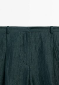 Pantalon en tissu texturé vert foncé avec passants pour ceinture, détails de couture à l'avant, et coupe ample visible de la taille jusqu'au milieu de la cuisse.