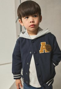 Veste varsity bleu marine avec capuche grise, ornée d'un écusson "A" crème et d'un accent en forme d'ours texturé. Poignets rayés et boutons.