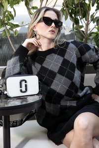 Maglione a scacchi bianco e grigio con scollo rotondo; borsa bianca con logo nero; modella che indossa occhiali da sole neri e orecchini a cerchio.