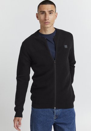 Svart glidelåsgenser med teksturert strikkedesign, ribbede mansjetter og en liten logo på brystet, slitt over en marineblå skjorte og denimjeans.