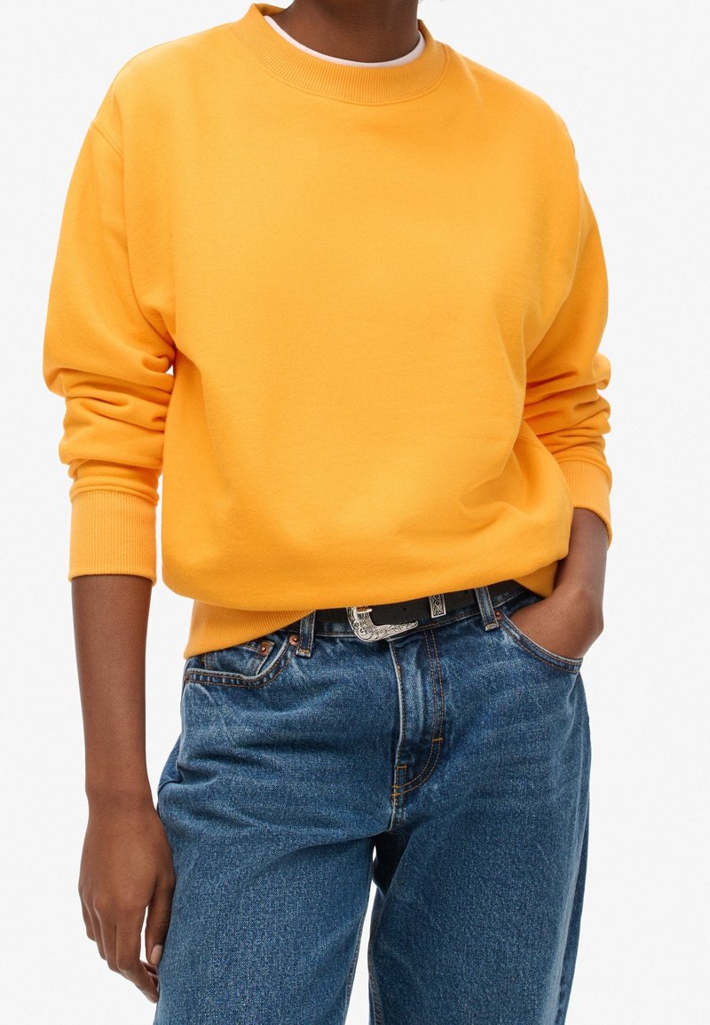Persona vestida con un sudadera de manga larga de color naranja brillante metida en unos jeans de tiro alto azules con un cinturón negro, con una mano en el bolsillo.
