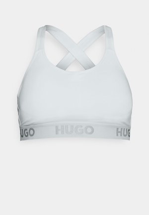 Soutien-gorge de sport gris clair en tissu lisse avec des bretelles croisées et une large bande portant le mot "HUGO" en lettres argentées.
