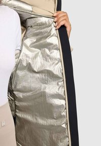 Prepínacia bunda s lesklou striebornou vnútornou podšívkou, krémovým vonkajším obalom a čiernym lemovaním. Má zipsové zatváranie a textúrované materiály.