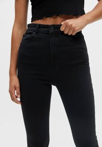 Persona vistiendo jeans negros de cintura alta y una blusa negra corta, con la mano metida en la cinturilla delantera, contra un fondo liso.