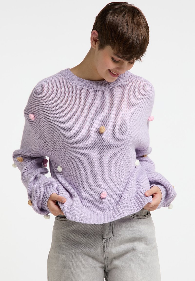 Lila gestrickter Pullover mit rundem Ausschnitt, verziert mit bunten Pompons und gerippten Bündchen. Kombiniert mit hellgrauen Jeans.