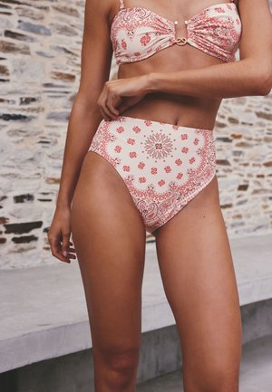 Femme portant un bas de bikini taille haute à motifs rouges et blancs avec un haut assorti sur un fond de mur en pierre