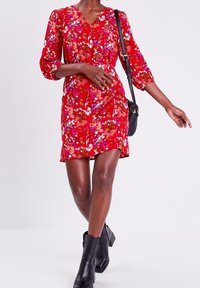 Robe rouge à fleurs avec des manches trois-quarts, un col en V et des détails froncés à la taille. Portée avec des bottines noires et un petit sac.