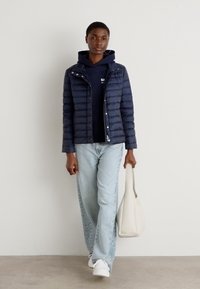 Giacca a vento blu navy con cuciture orizzontali, indossata sopra un'hoodie scura. Jeans azzurri e sneaker bianche. Borsa beige in mano.