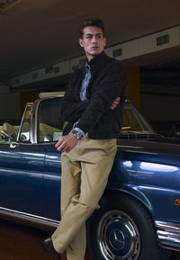 Uomo vestito con una giacca scura, camicia a quadri e pantaloni beige si appoggia a un'auto cabriolet vintage blu con accenti cromati.