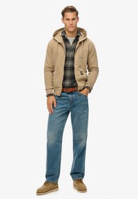Sudadera beige con cremallera y cordones sobre un cuello redondo gris, camisa a cuadros, jeans azules y calzado beige. Diseño simple, ajuste casual.