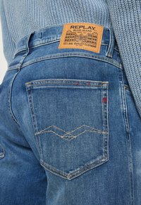 Niebieskie dżinsy z denimu z skórzaną metką, posiadające przeszywany wzór na tylniej kieszeni oraz czerwone akcentujące przeszycia. Teksturowana tkanina.