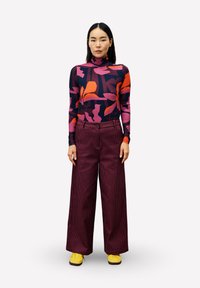 Langarm-Turtleneck mit auffälligen bunten Blumenmustern, kombiniert mit weiten burgunderroten Cordhosen und gelben Sneakern.