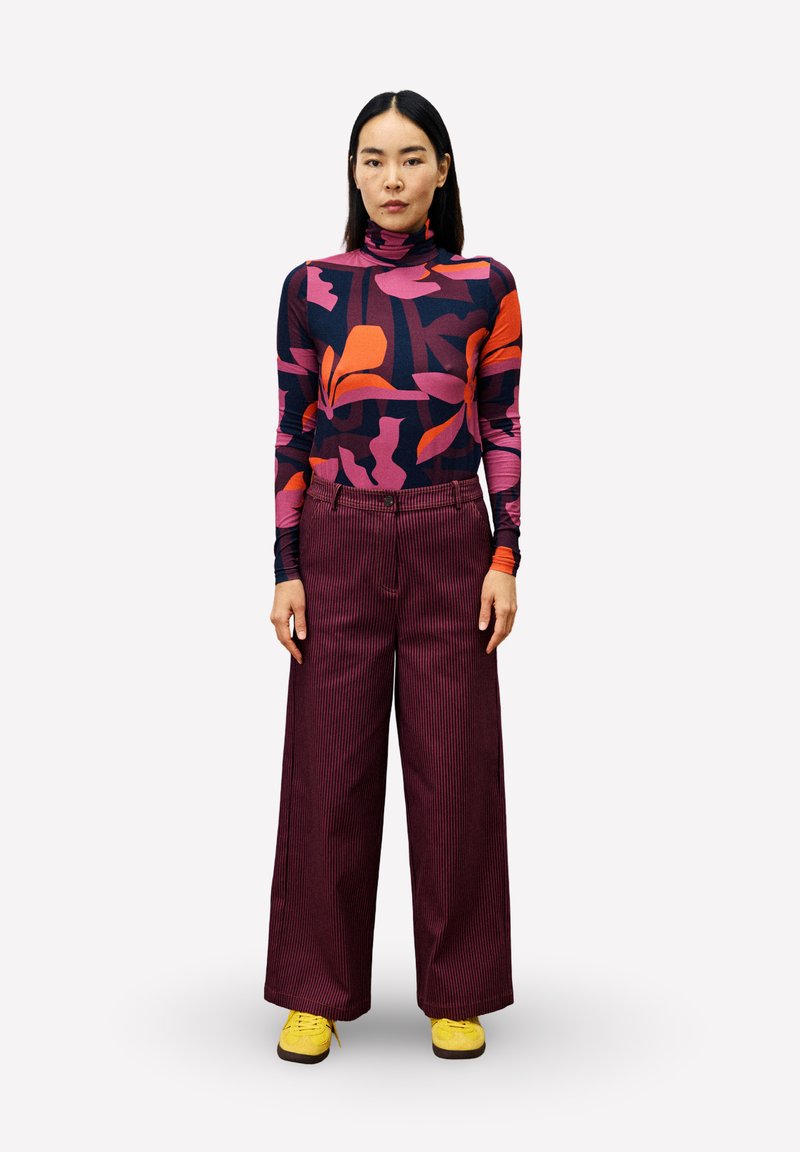 Langarm-Turtleneck mit auffälligen bunten Blumenmustern, kombiniert mit weiten burgunderroten Cordhosen und gelben Sneakern.